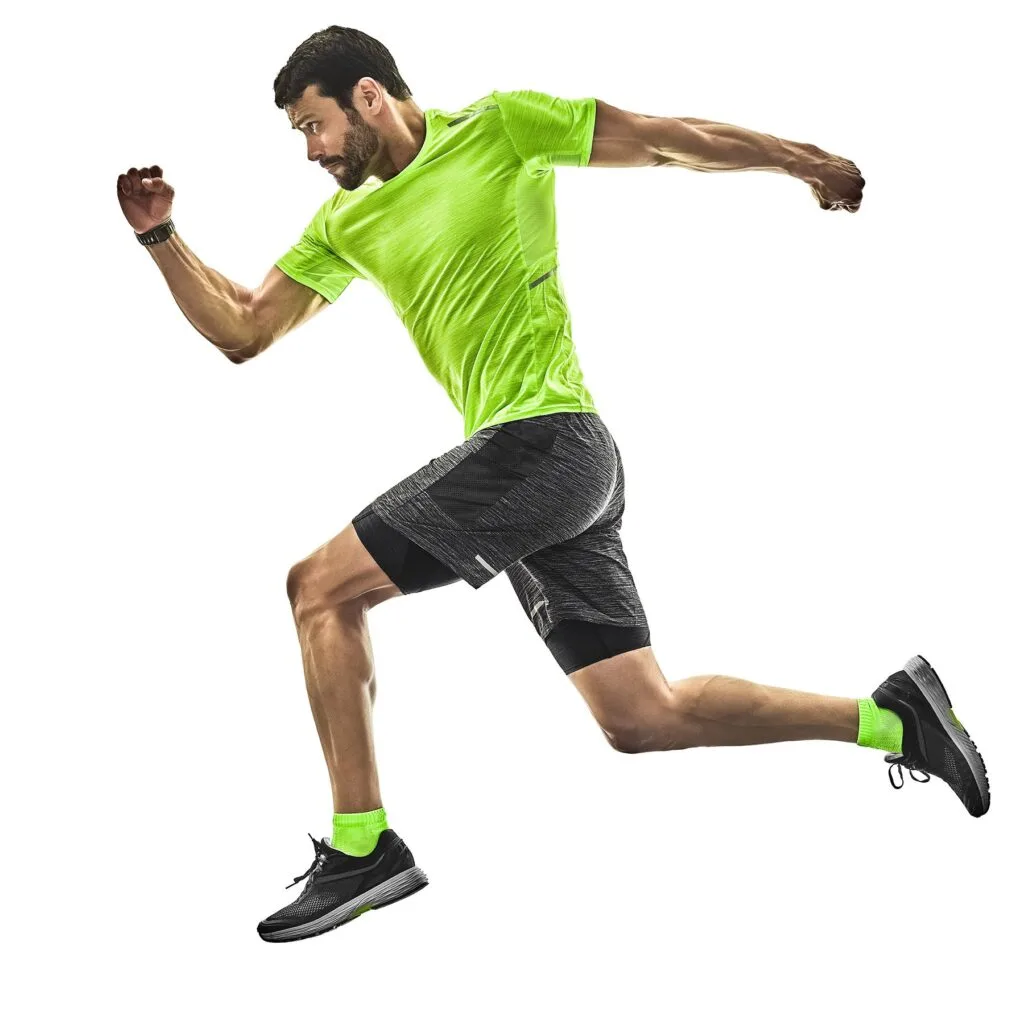 A man in a green shirt and black shorts runs energetically towards a pickleball court, ready to join the game. The background features a vibrant outdoor setting with trees and a clear sky, emphasizing the excitement of the sport.